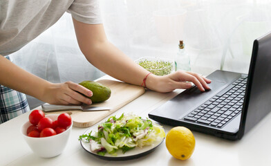 Obraz na płótnie Canvas A woman holds an avocado in her hand and looks for a recipe for a vegan salad on her laptop. Lettuce and ingredients are on the table. Online cooking concept. Horizontal orientation.