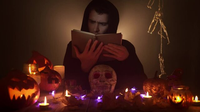 Portrait of man in the image of wizard reading voodoo book among candles and jack o'lanterns on black background. Scary wizard is practicing witchcraft over a skull surrounded by Halloween decorations