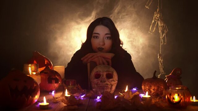 A bored young woman in the image of witch is sitting leaning on the magic book and knocking fingers on the ritual skull. A pretty witch in a dark gothic costume is surrounded by Halloween decorations.