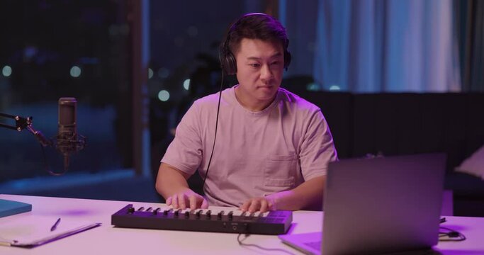 Asian talented young male artist recording music tracks on electronic piano keyboard synthesizer enjoying rhythm playing audiotracks in headphones listening. Home lifestyle.