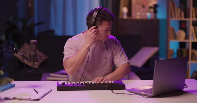 Home lifestyle. Chinese young adult male in positive mood creating new music generating ideas making music recording audiotrack listening in headphones. Electronic piano.