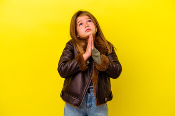 Little caucasian girl isolated on yellow background holding hands in pray near mouth, feels...