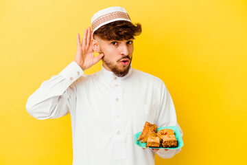 Young Moroccan man wearing the typical arabic costume eating Arabian sweets isolated on yellow background trying to listening a gossip.