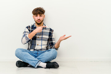 Young Moroccan man sitting on the floor isolated on white background excited holding a copy space on palm.