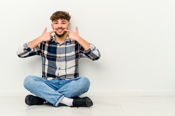 Young Moroccan man sitting on the floor isolated on white background smiles, pointing fingers at mouth.