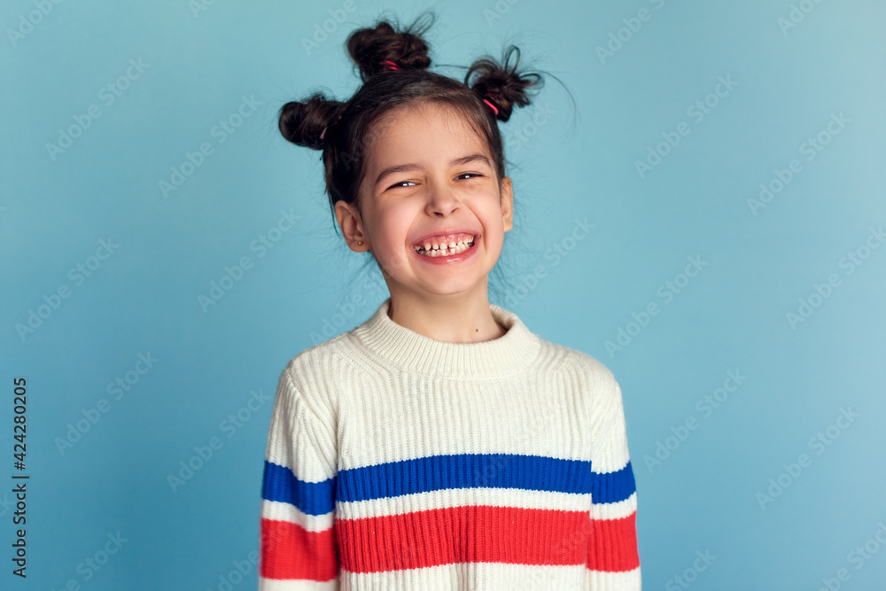 Wall mural Adorable little girl with funny hairstyle, smiling broadly isolated over studio blue color background. Playful kid has a happy expression on her face.