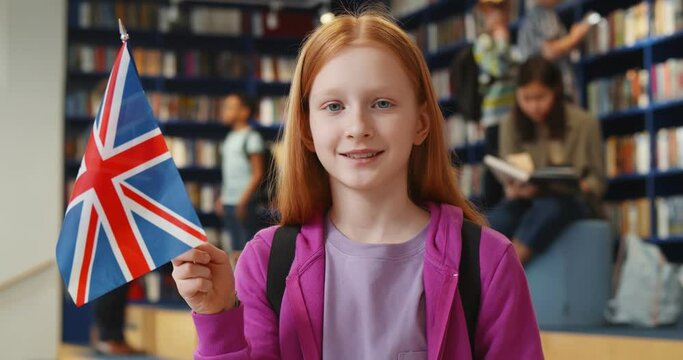 Pretty preteen girl holding great britain flag and smiling at camera standing in school library