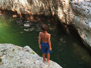 A boy in blue shorts stands on the edge of a canyon in front of a river flowing into the gorge