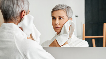 Mature woman with short grey hair looking at her reflection in bathroom mirror while wiping cheek - Powered by Adobe