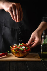 The chef cook sprinkles salted fresh vegetable salad in plate on a wooden table. Cooking healthy food in the kitchen in a restaurant