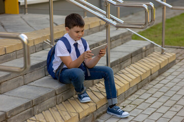 Schoolboy brunette in a white shirt with a blue jacket sits on the stairs and plays with a gray tablet.