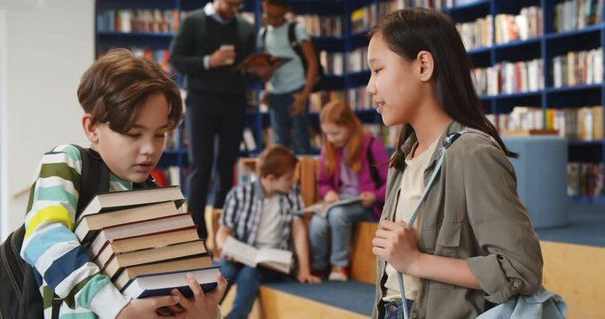 Schoolboy holding stack of books and talking to classmate in library