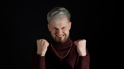 Portrait of overjoyed young man in trendy stylish clothes wins competition achieves goal and makes triumph gesture, guy says yes feels like winner shows fist, happy dream come true on black background