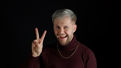 Friendly happy stylish young man with blue eyes in trendy clothes looking at camera, feeling happiness, fun, enjoyment, showing victory sign, hoping for success and win, doing peace gesture, smiling