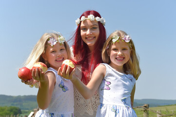 A beautiful young woman wearing white vintage
clothing and twin sisters stand together.They show
off their apples they are eating. On their faces is an 
expression of happiness.