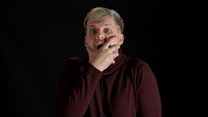 Fototapeta premium Shocked amazed handsome man in stylish blouse feeling excitement, looking at camera and holding hands near face on black background. Facial expression of trendy bearded guy surprised say wow omg