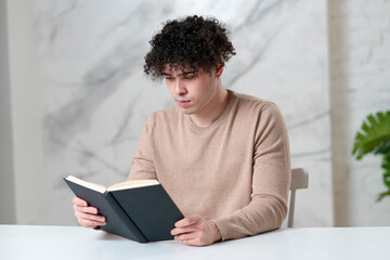 Portrait of handsome ethnic serious guy student, young concentrated intelligent man reader is reading an interesting book with interest at home at table, studying. Literature, leisure concept.
