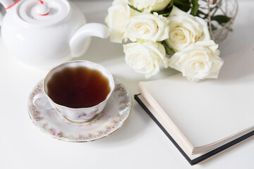 A porcelain tea cup with a saucer, a teapot, a white faience angel and three roses on the table. Breakfast.