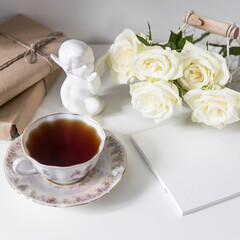 A porcelain tea cup with a saucer, a teapot, a white faience angel and three roses on the table. Breakfast.