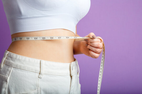 Slim Woman Measures Her Waist Waistline With Measuring Tape. Healthy Body Shaping Weight Loss Concept. Slim Waist Small Belly In Big White Denim Pants Isolated Over Purple Color Background. Copy Space