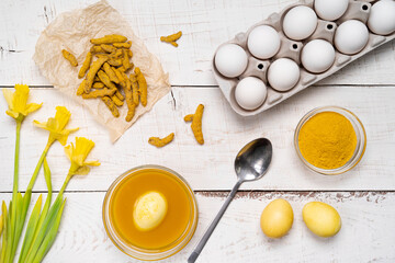 the process of painting Easter eggs with natural plant dyes, turmeric, on a white wooden background, top view of chicken eggs and yellow daffodils