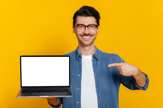 Satisfied confident caucasian unshaven stylish guy with glasses, holds an open laptop with blank white screen, points finger at it, standing on isolated orange background, smiling friendly