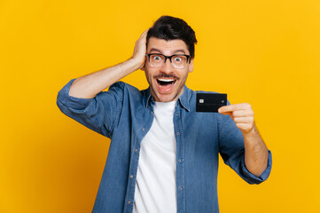 Happy cheerful stylish handsome caucasian guy with glasses, holds a credit card in hand, touches...