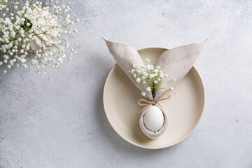 Easter table setting with egg in napkin in the form of an Easter bunny with ears, with small white flowers on ceramic plate. Easter table decorations. Top view, copy space .