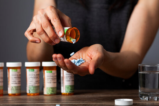 a young depressed woman is taking pills out of the medication bottle. She has a stack of bottles lined up at the back. Concept image for drug use, over dose, prescription medication, depression
