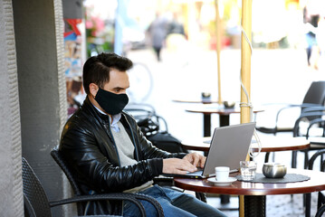 Man with protective mask for Covid-19 virus working on laptop in a public bar. University student  sitting on a bench and using computer in a public place to access internet or social media.