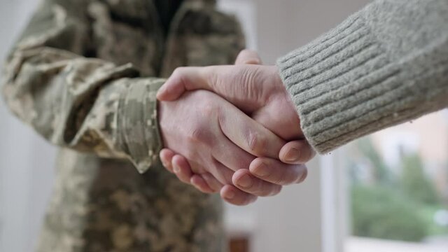 Close-up handshake of middle aged soldier in camouflage and young Caucasian man. Unrecognizable military father and adult son shaking hands indoors at home. Unity and family support