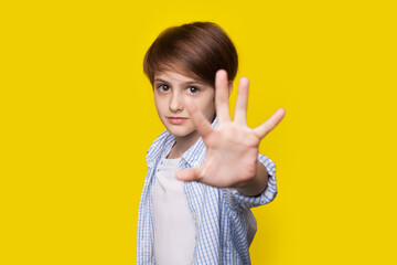 Caucasian small boy is gesturing with palm the stop sign posing on a yellow studio wall