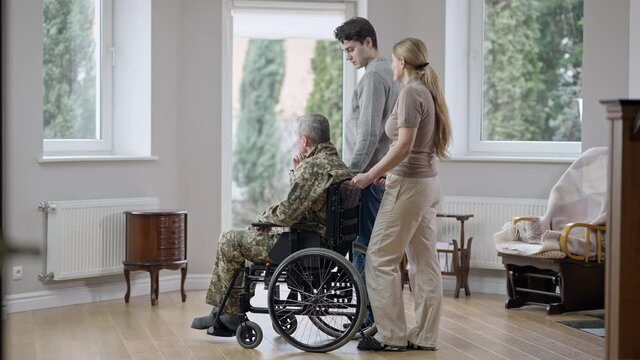 Side view wide shot of Caucasian family looking out the window talking. Disabled paraplegic military man, wife and son chatting at home indoors. Family support and unity concept