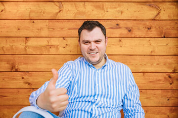 Young man in striped shirt sits next to a wooden wall, showing thumbs. Have a nice vacation. Concept of happiness and contentment 