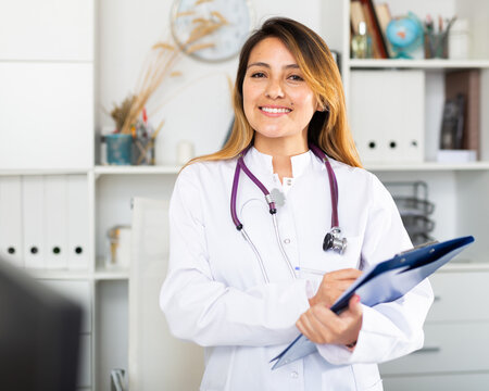 Young latina woman doctor assistant standing in medical office making notes on clipboard