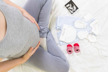 Top view of expecting pregnant woman in the bed with ultrasound scans and preparing baby clothes at home, boy or girl, pregnancy and birth concept