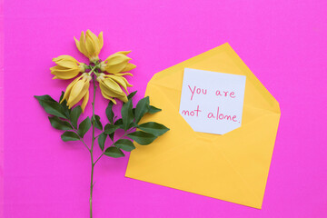 you are not alone message card handwriting  with yellow flowers ylang ylang in envelope arrangement flat lay postcard style on background pink