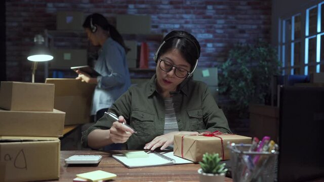 two young Asian casual women colleagues working small business online packing talking to clients by headsets and note orders on customers boxes. internet shop female sellers in night working.