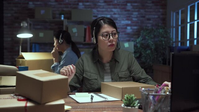 Young asian woman seller talking phone call with customer for online business on headset. girl staff discussing and helping client solving problem with parcels. lady coworker overtime working night