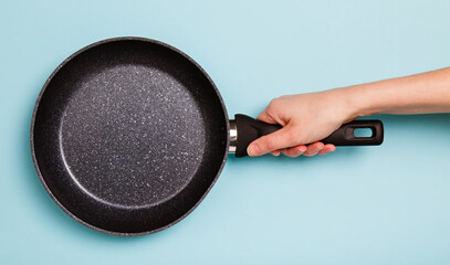 frying pan in woman's hand on blue background. Woman holding a pan in a hand. Female hand holding simple new empty Non-stick Frying Pan with black handle