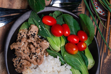 Teriyaki pork sprinkled with sesame and vegetables and steamed rice on a black plate.