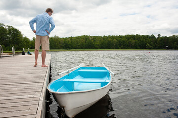 A man standing on a wooden pier looks at a boat with oars in the water.