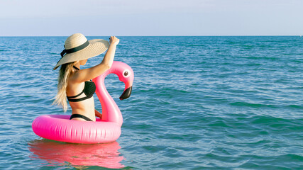 Summer beach relaxing. Happy young sexy girl in bikini swimsuit, sunglasses and straw hat with pink inflatable flamingo in blue sea water on ocean background. Trendy summer concept.