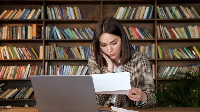 Young woman with short dark hair opens bank letter and touches cheek with shock sitting at laptop against wooden bookshelves at home