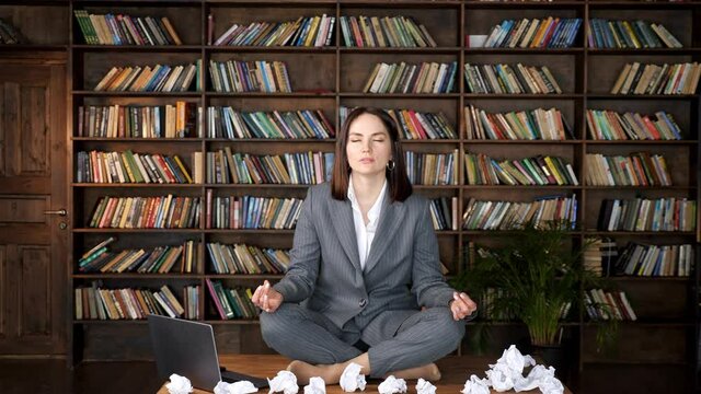 Short haired businesswoman meditates sitting in yoga pose on wooden table among rumpled paper sheets and laptop against bookshelves