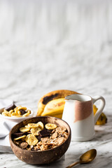 Muesli made from mix of unprocessed whole grains, chia, quinoa, nuts, seeds, with banana and chocolate in coconut vowl on the marble table . Heathy breakfast concept.