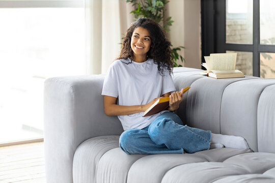 Happy woman at home sitting on the couch sitting on the couch reading book. Beautiful African American young woman relaxes reading book at home on sofa