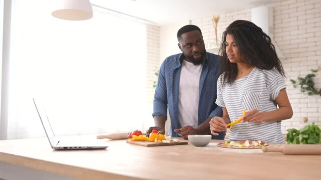 Excited young African American couple following steps from watching online cooking classes, learning how to make pizza, watching video blog course from laptop in the kitchen, placing vegetables