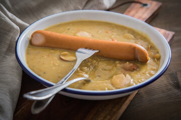 Deftige Deutsche Erbsensuppe als Eintopf mit Bockwurst in Emaille Teller auf Holzbrett und Holz  Hintergrund dunkel