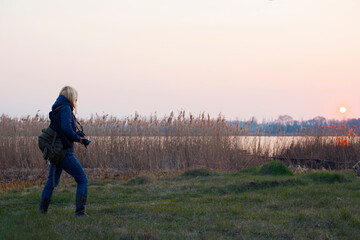 Fototapeta na wymiar girl photographer on the river bank blonde back view. traveler photographer, blogger, river bank overgrown with grass, setting sun. the concept of tourism, travel. autumn nature